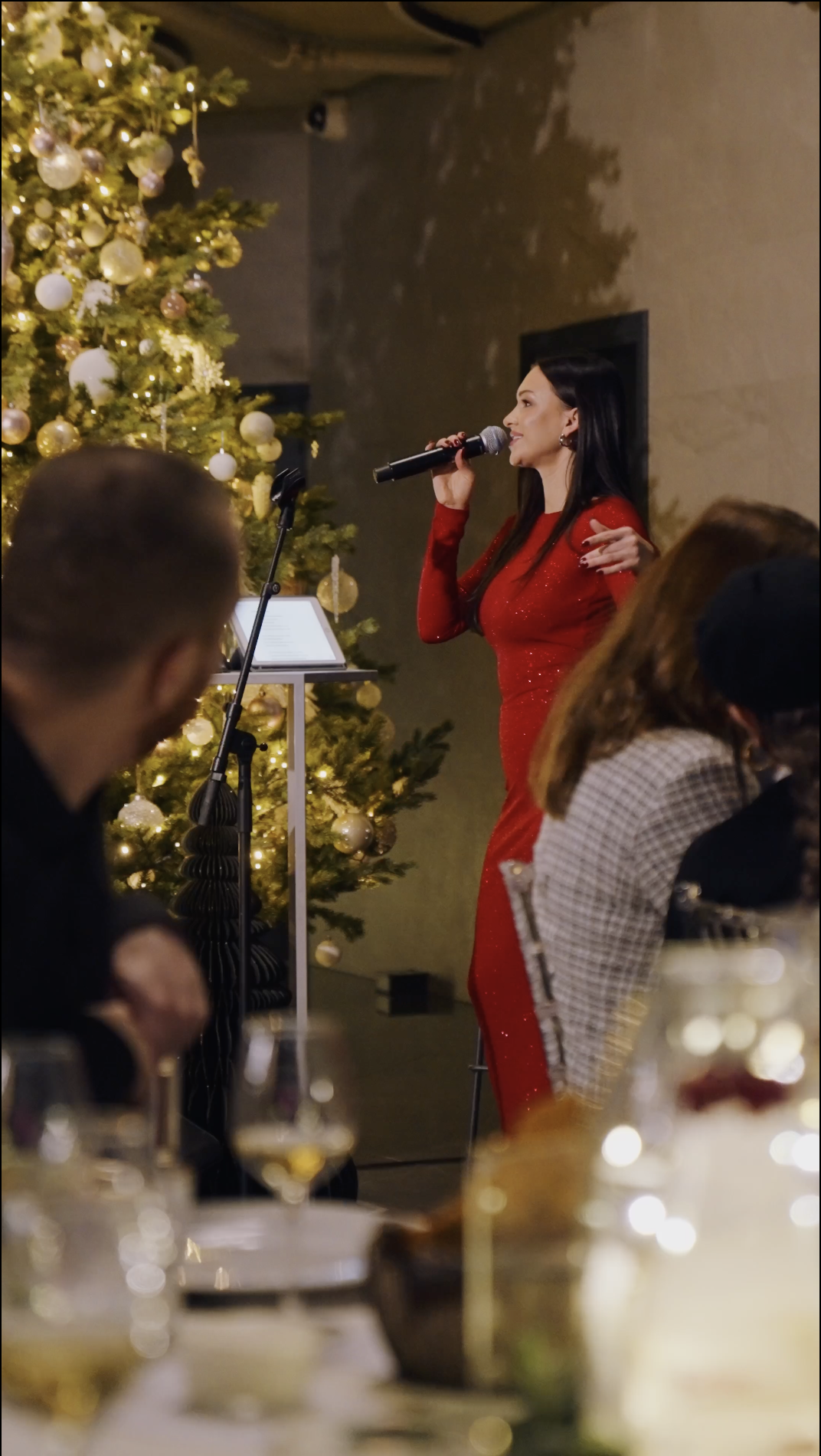 Host in a red dress speaking into a microphone beside a decorated Christmas tree at a festive dinner